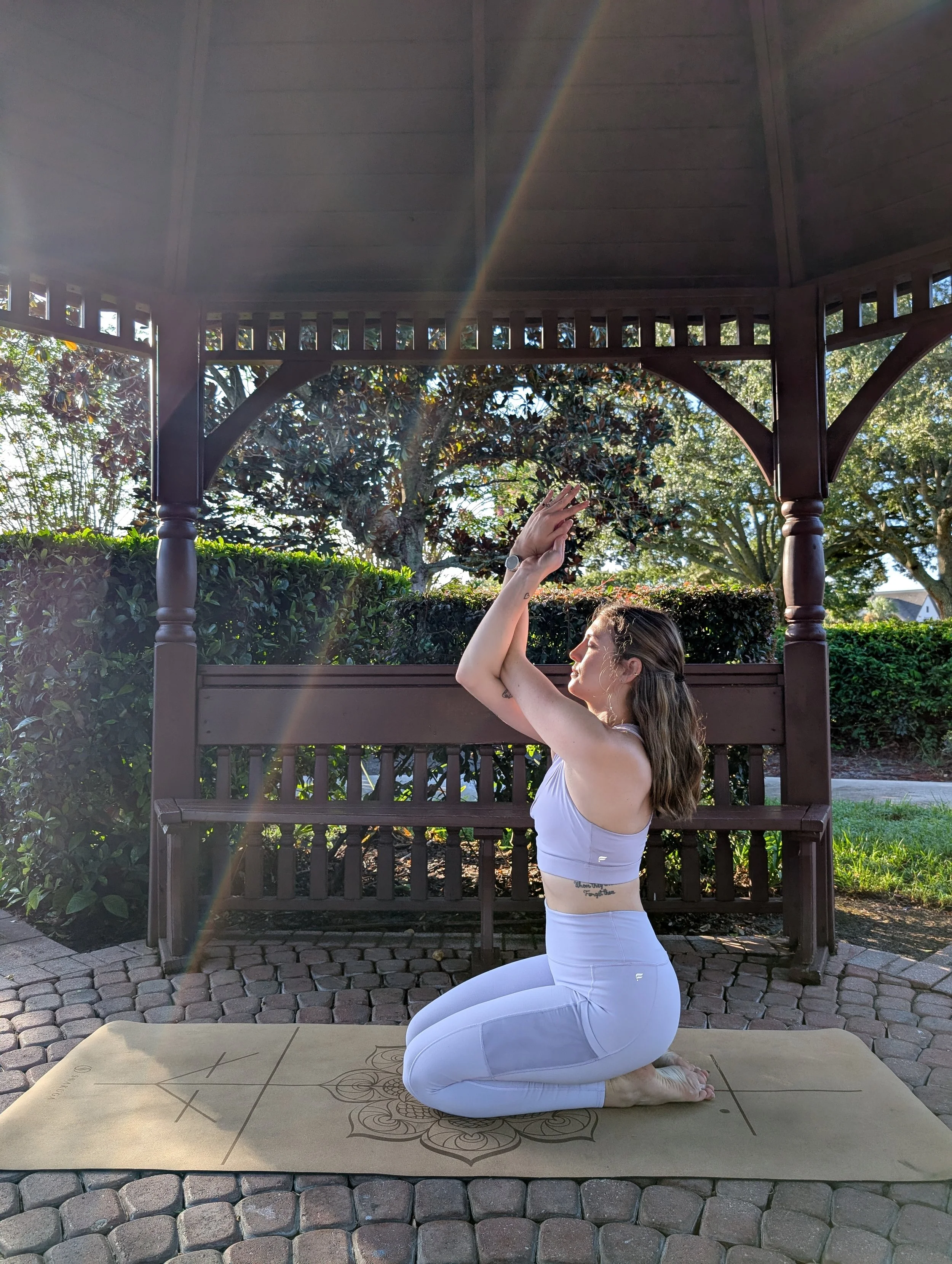 A woman practicing yoga outdoors on a yoga mat in a kneeling pose, with one arm raised and hand reaching toward the sky, under a wooden gazebo with trees and bushes in the background.