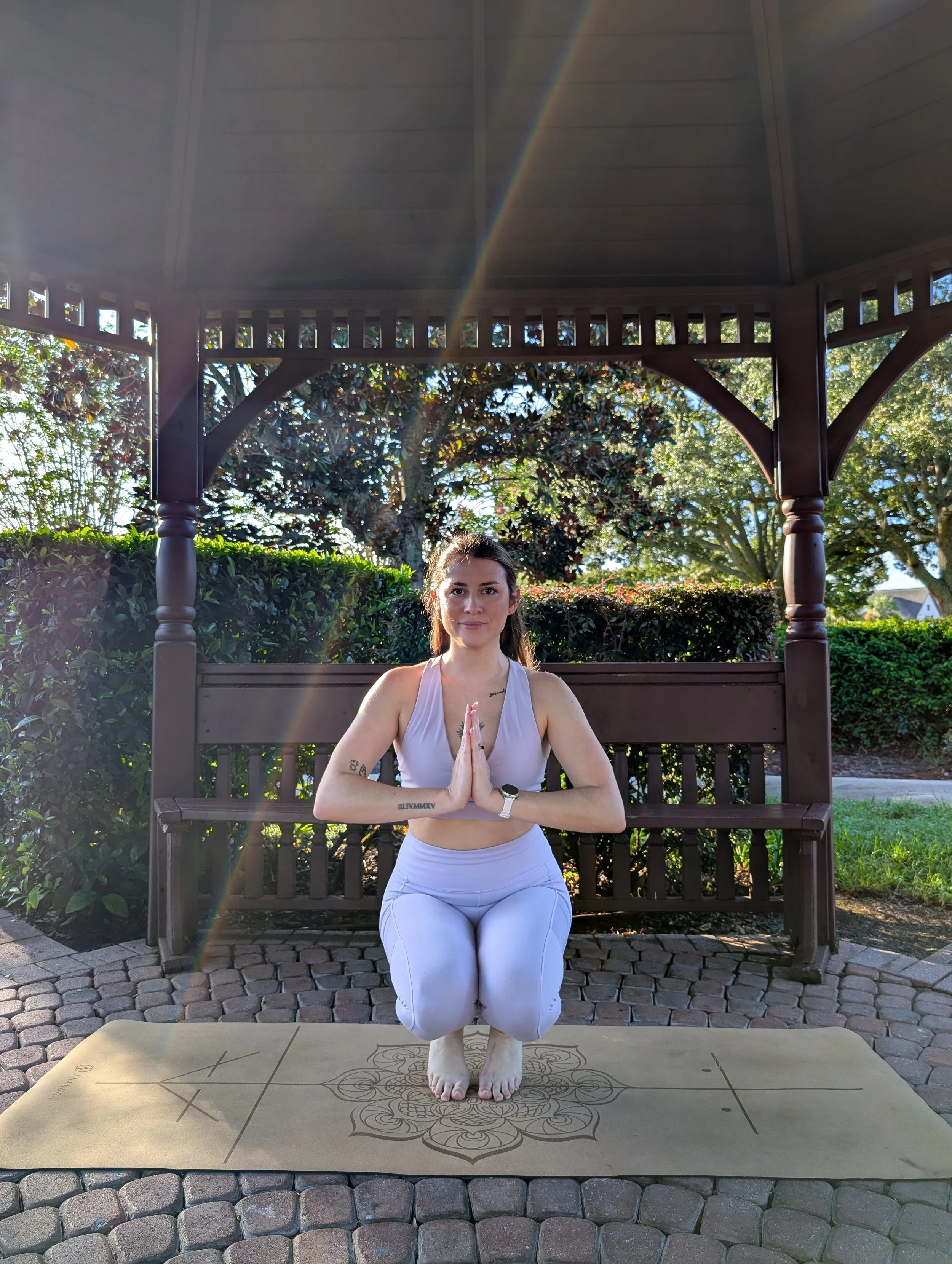 A woman practicing yoga outdoors on a tan mat with a floral design, sitting in a kneeling pose with hands in prayer position, under a wooden pavilion with trees and bushes in the background.