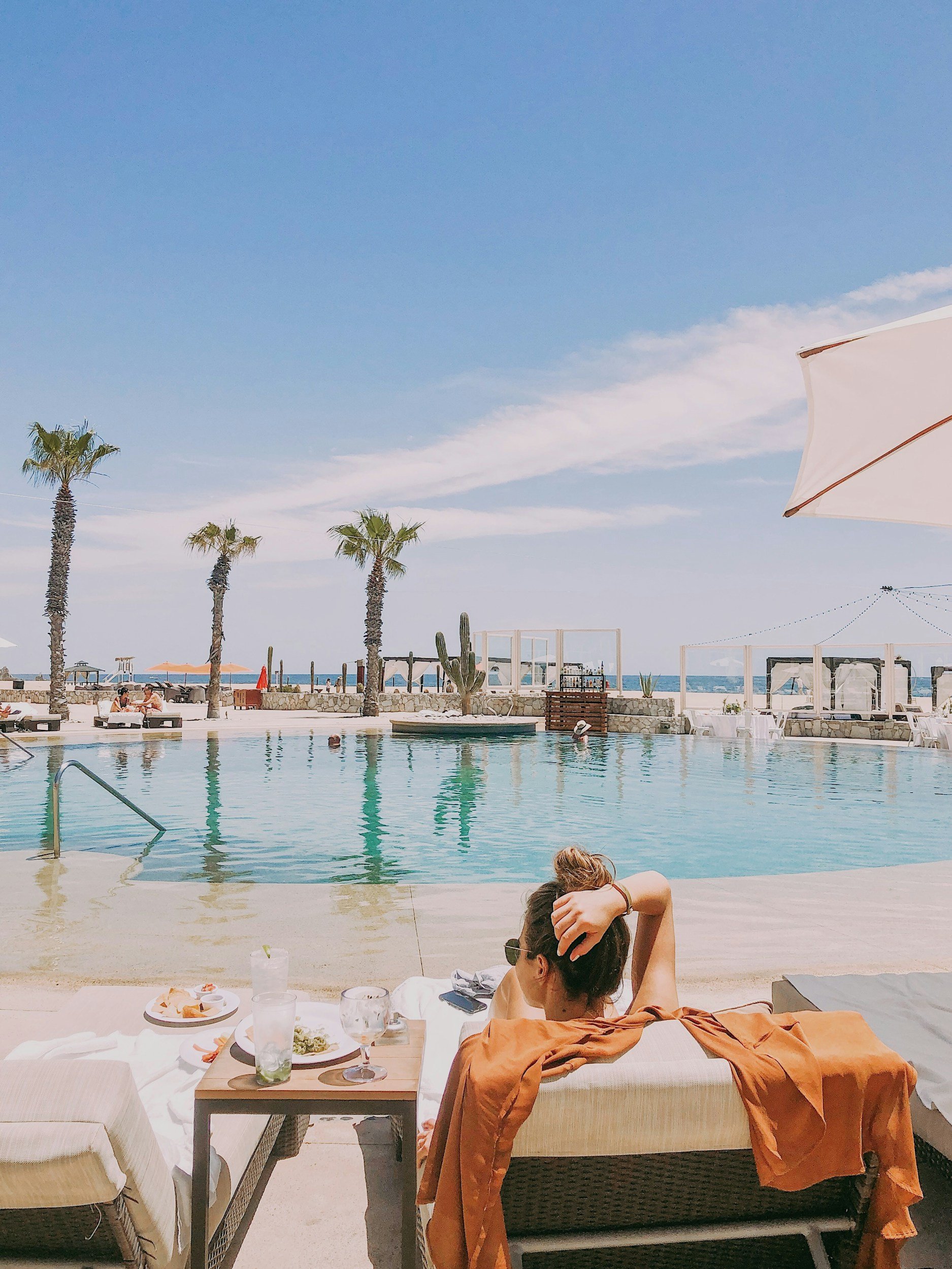 A woman is relaxing on a lounge chair at a beachfront poolside restaurant, with a table of food and drinks in front of her. There are palm trees, a swimming pool, and the beach with umbrellas and lounge chairs in the background, under a partly cloudy sky.