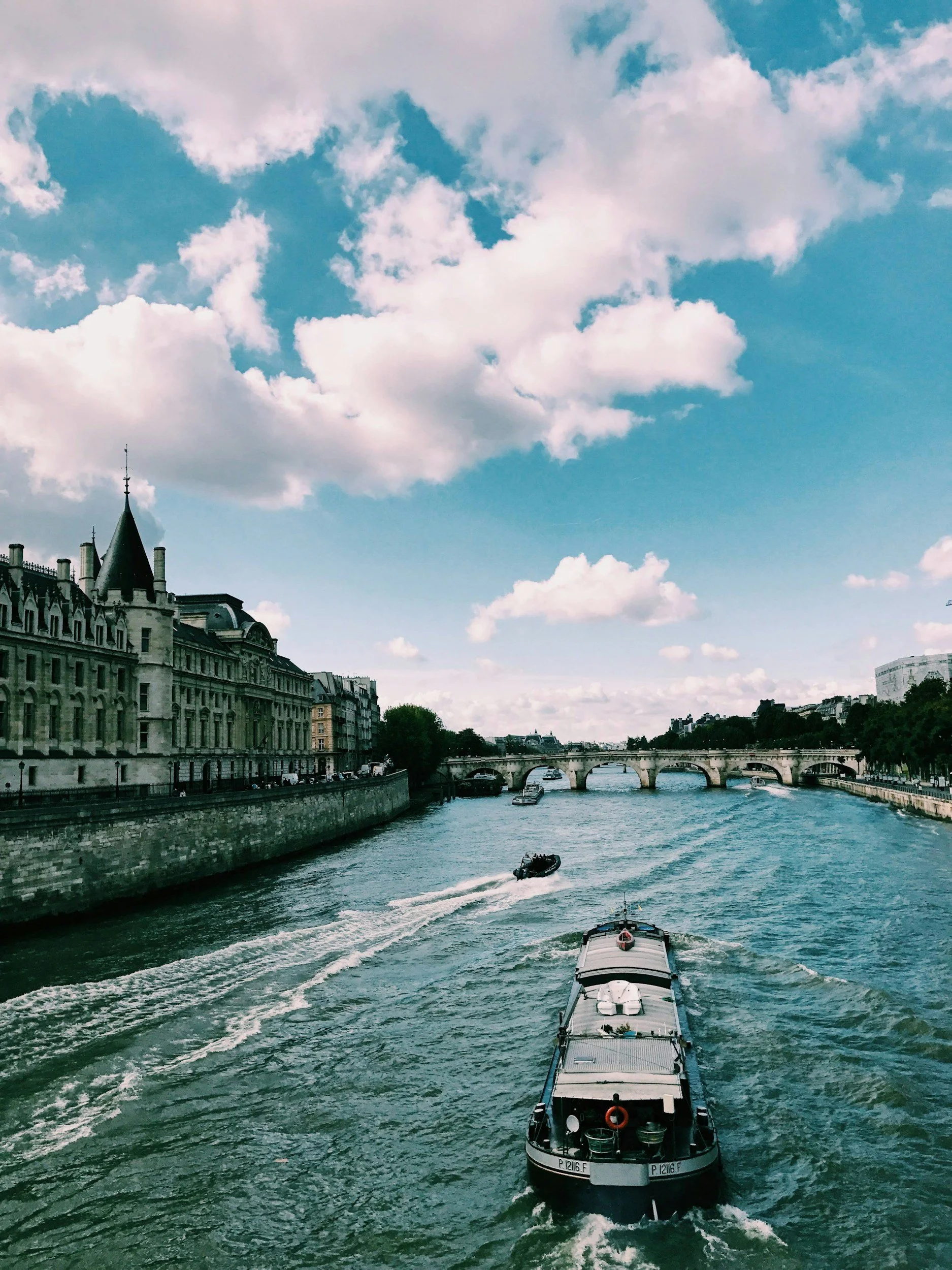 View of a river with boats and a bridge in the background, surrounded by historic buildings and a blue sky with white clouds.