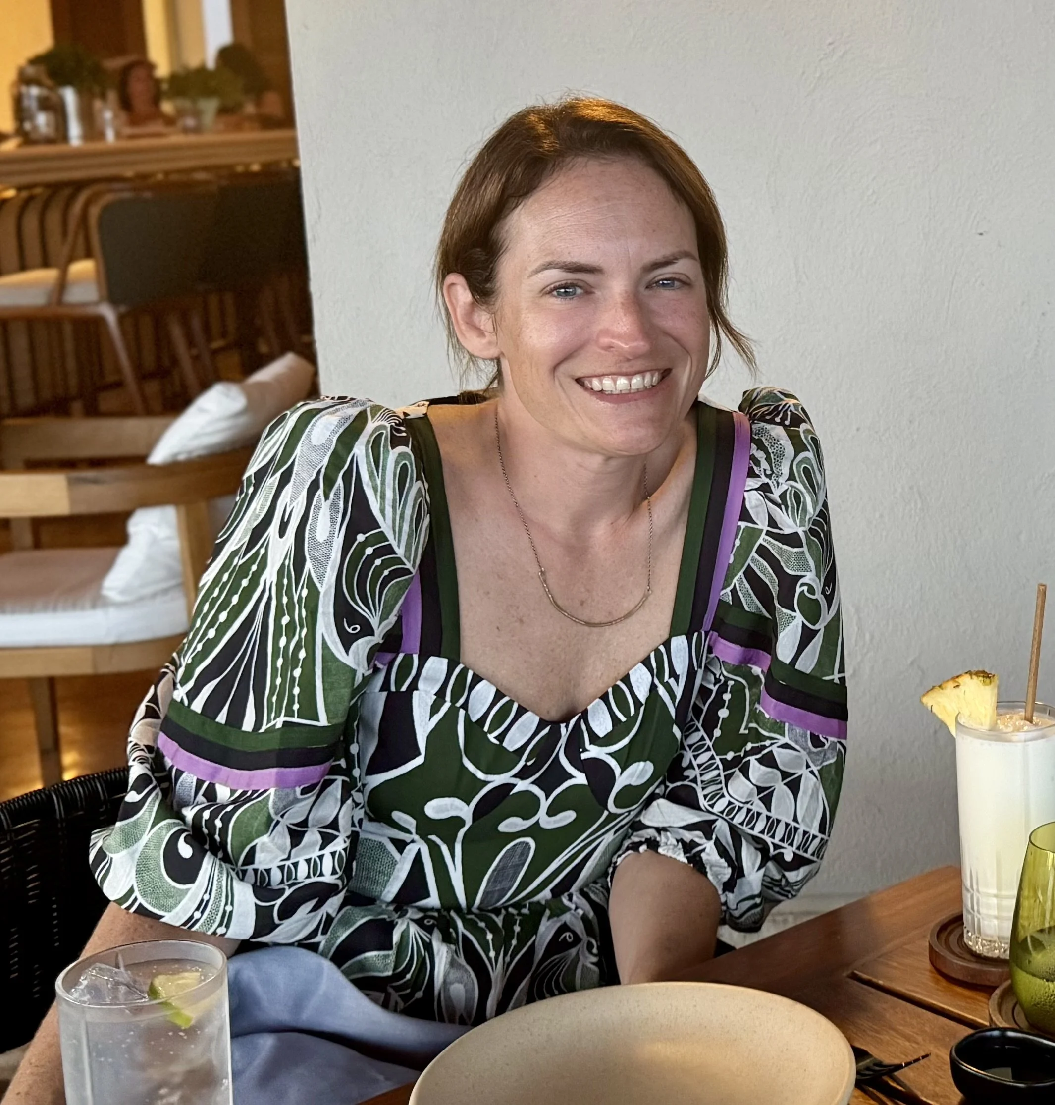 A woman with short red hair and blue eyes smiling, wearing a patterned top with black, green, white, and purple colors, sitting at a wooden table with drinks, plates, and napkins in a restaurant or cafe, with a white wall in the background.