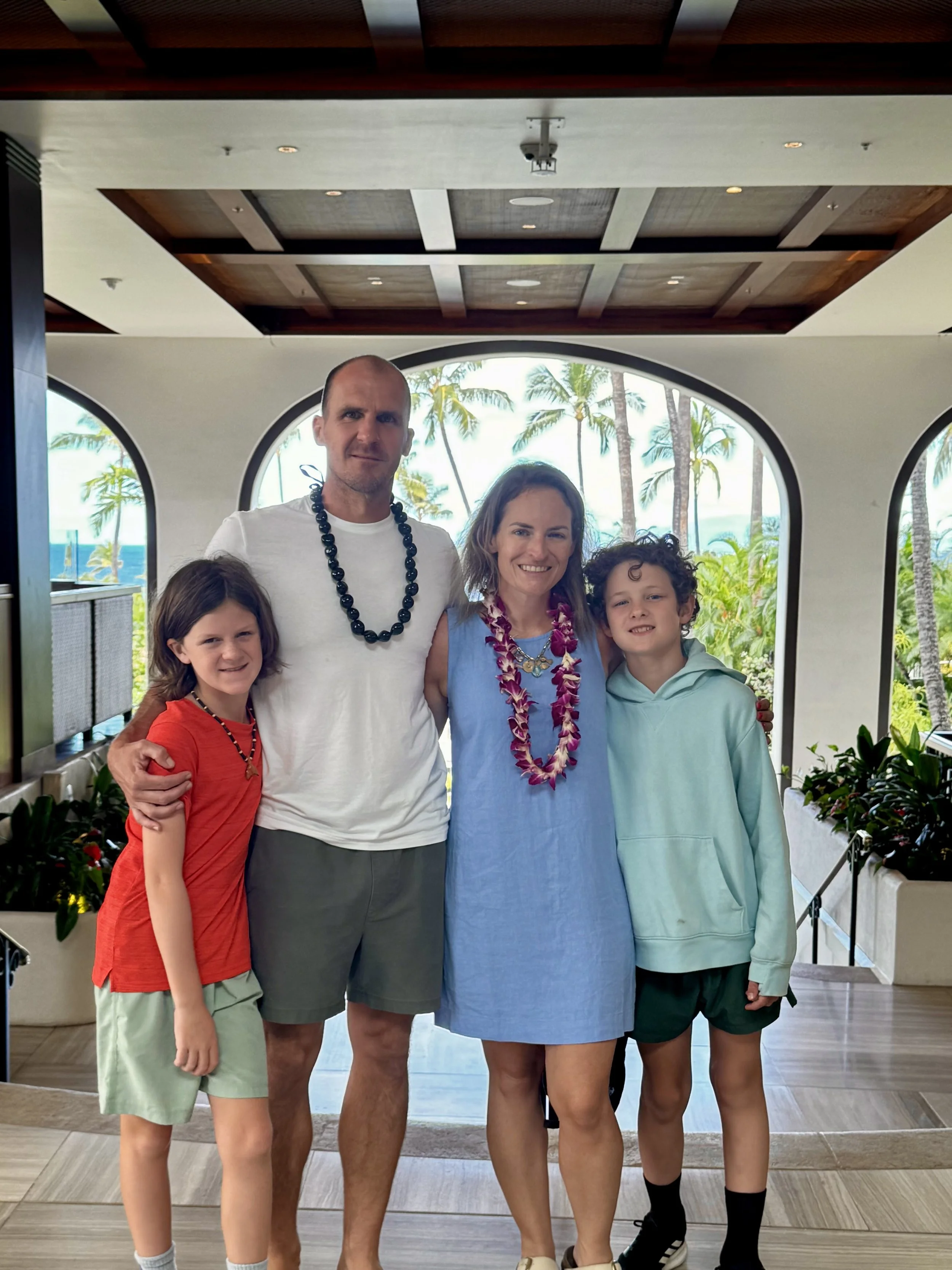 A group of four people, two adults and two children, standing inside a tropical-themed hotel or resort with palm trees visible through large arched windows in the background.