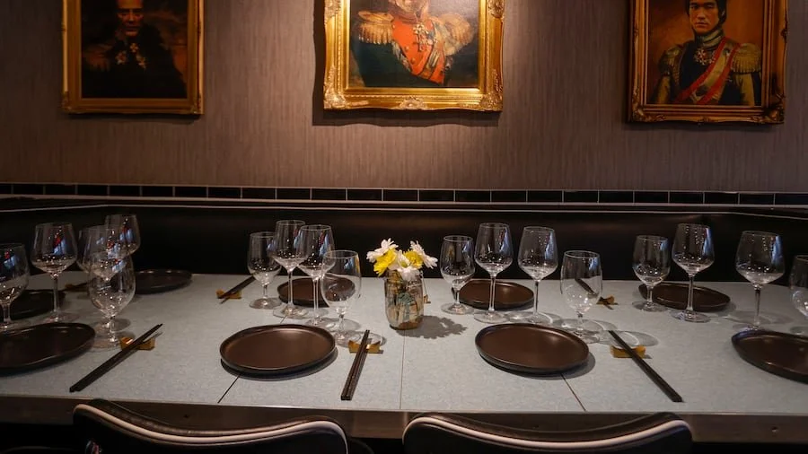 A dining table set with multiple wine glasses, brown plates, black chopsticks, and a small vase with yellow and white flowers, in a room with framed portraits on the wall.