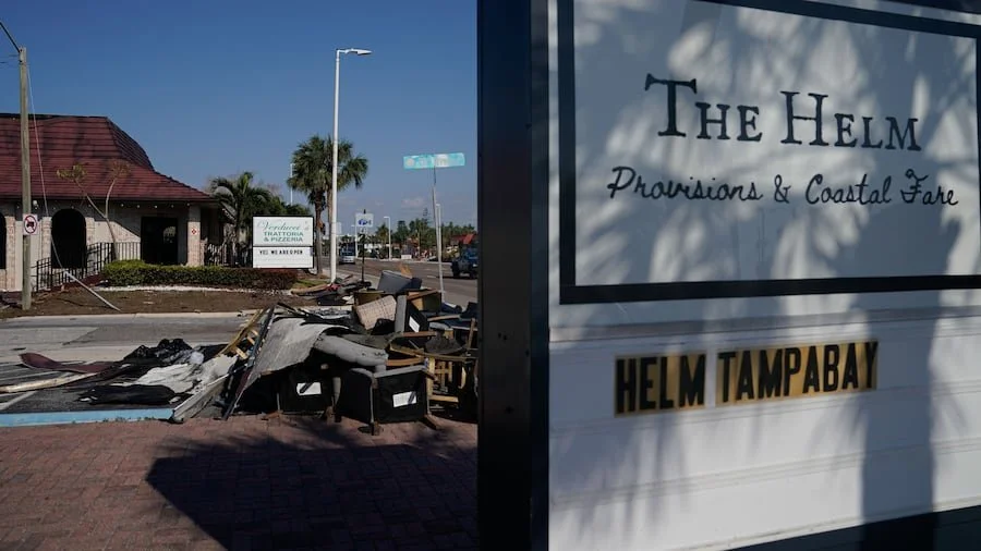 A large sign for The Helm Provisions & Coastal Fare with yellow and black text reading 'Helm Tampabay' in front of a pile of debris and garbage on the sidewalk, a building with a red roof and palm trees in the background.
