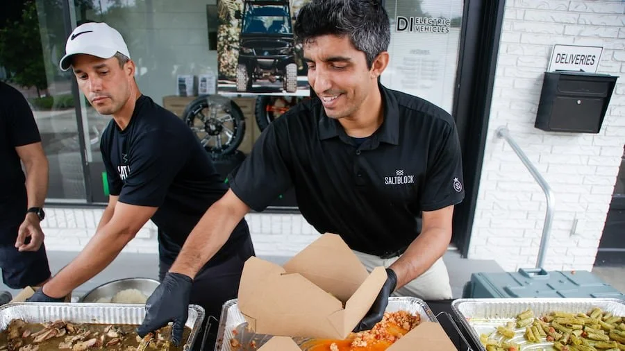 Two men serve food at an outdoor event, one smiling and placing food into a box, the other looking down. Food trays with meat, vegetables, and side dishes are in front of them, and they are wearing black shirts and gloves.