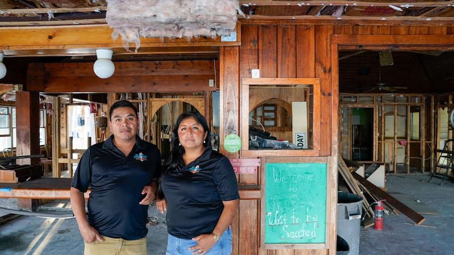 Two people standing inside a partially constructed building with wooden framing, insulation, and construction materials around them.