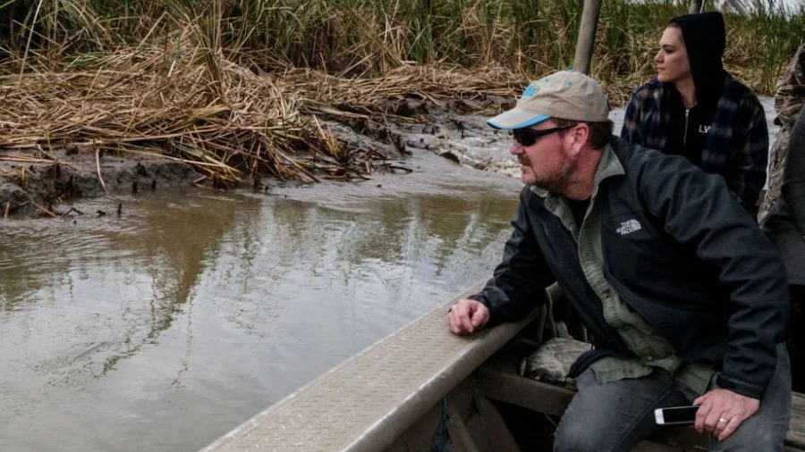 Two people on a boat observing a muddy river, with grass and shrubs on the riverbank.