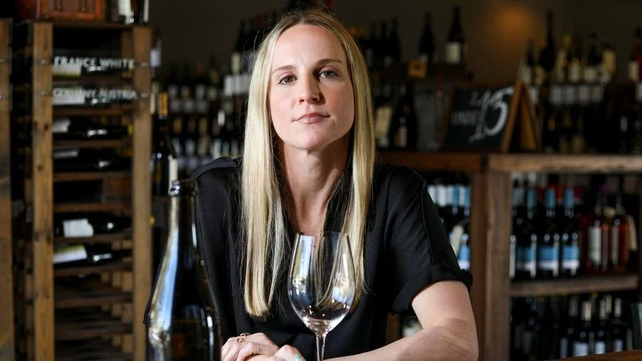A woman with long blonde hair sitting at a table in a wine shop or bar, with bottles of wine on shelves behind her, and a wine glass in front of her.
