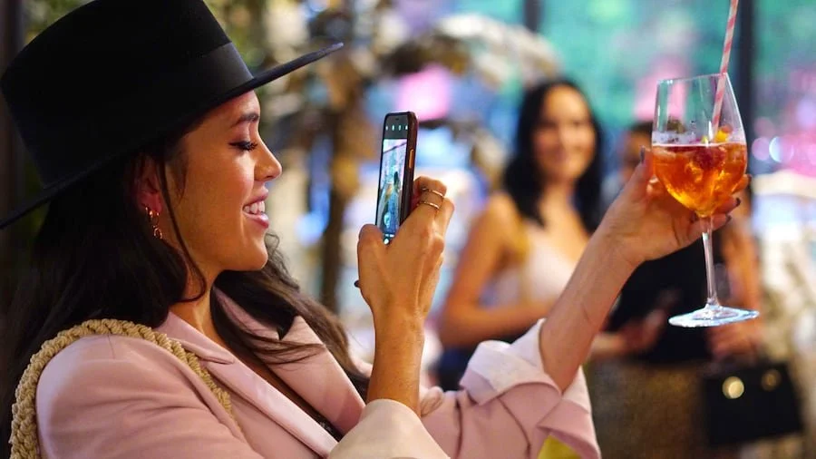 Woman wearing a black hat takes a photo of her colorful cocktail with a straw using her smartphone, while smiling at a social gathering.