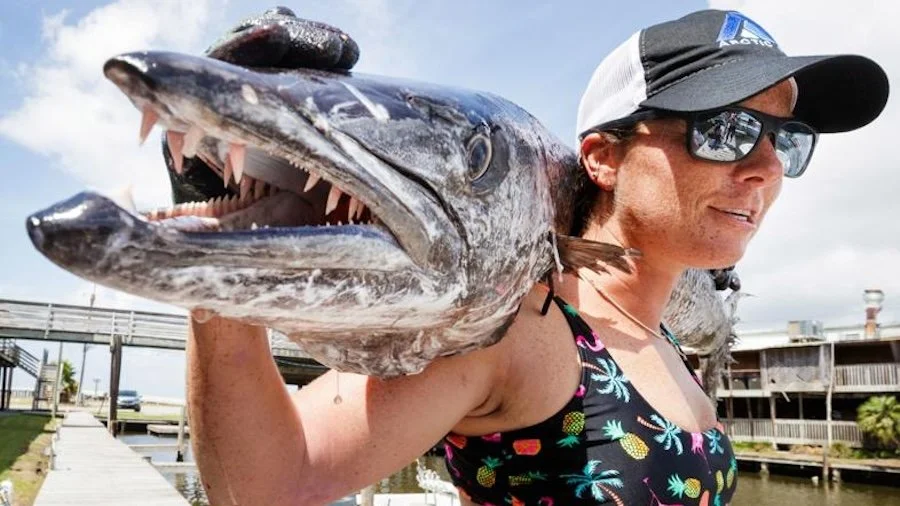 Woman wearing sunglasses and a cap, holding a large fish over her shoulder near a marina, with boats and a walkway in the background.