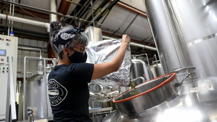 A woman wearing a black mask and black bandana with a white pattern, pouring ingredients into a large stainless steel brewing tank in a brewery.