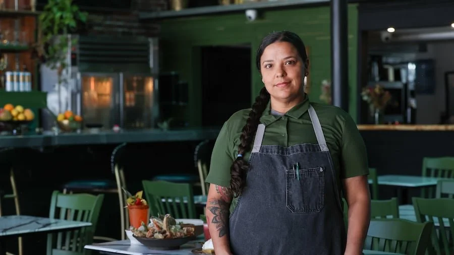 A woman with a braid wearing a green shirt and a black apron standing in a restaurant or cafe, with tables, chairs, and a bar area in the background