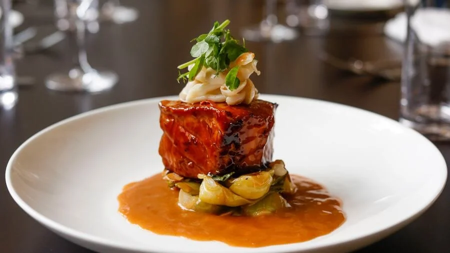 A plated gourmet dish featuring a glazed piece of meat atop a bed of vegetables, with sauce, garnished with pasta and microgreens, on a white plate.