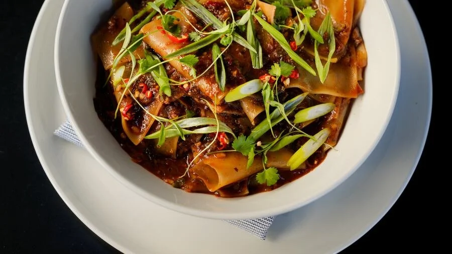 A bowl of Asian-style noodle dish with wide rice noodles, beef, and chopped vegetables, garnished with chopped green onions, cilantro, and microgreens.