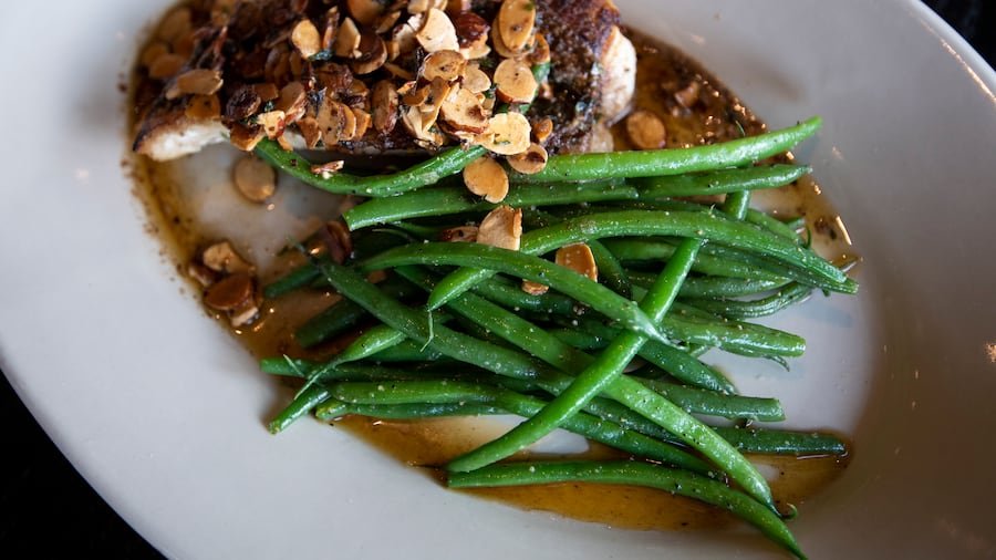 Plate of cooked green beans with chopped nuts and a piece of meat topped with chopped nuts, served with brown sauce.