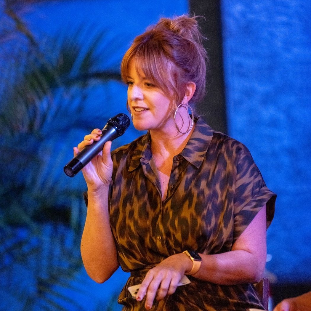 A woman with pink hair styled in a messy bun, wearing a leopard print blouse and hoop earrings, is holding a microphone and speaking on a stage with a blue background and palm leaves.