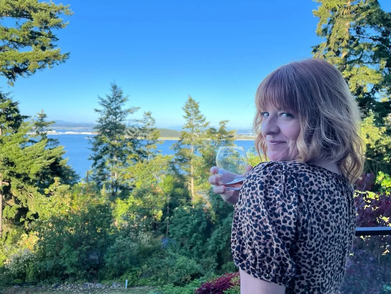 A woman with shoulder-length blonde hair and a leopard print top holding a wine glass, standing outdoors with a view of trees, water, and a blue sky in the background.