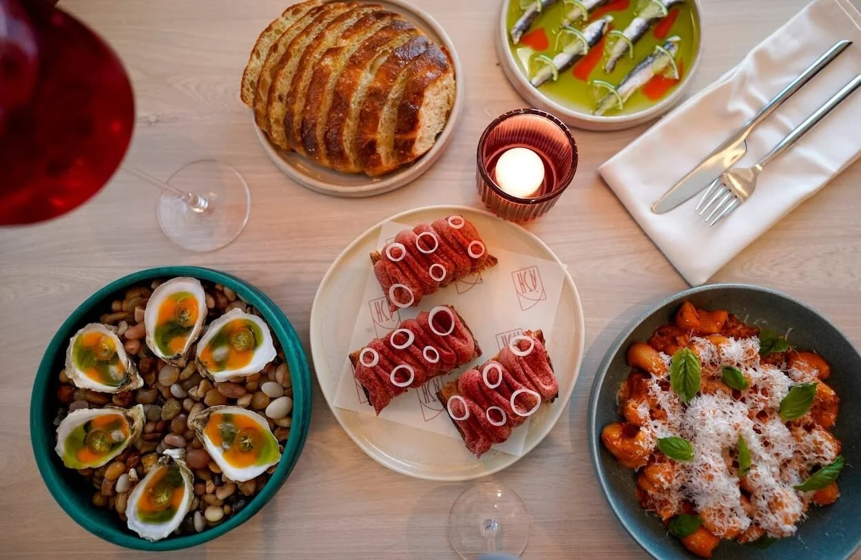 A table set with various Spanish dishes including sliced bread, green olives with red pepper, slices of cured ham garnished with rings of onion, a bowl of roasted vegetables topped with grated cheese and basil, a bowl of beans with soft-boiled eggs, and a plate with small fish wrapped in olive oil with red pepper.