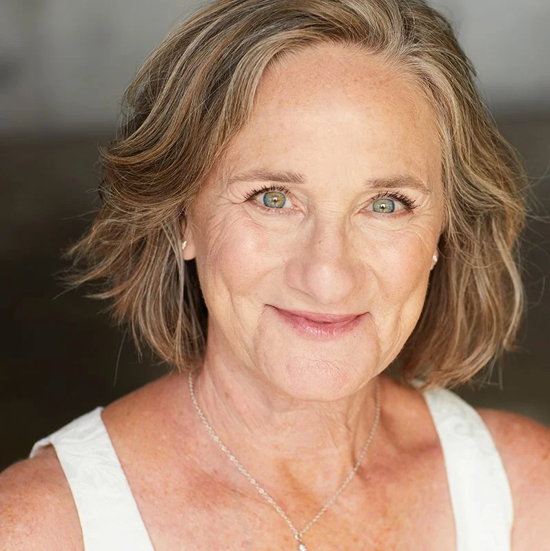 Lori Aronsohn. Close-up of an older woman with shoulder-length brown hair, smiling, wearing a white top and a silver necklace.