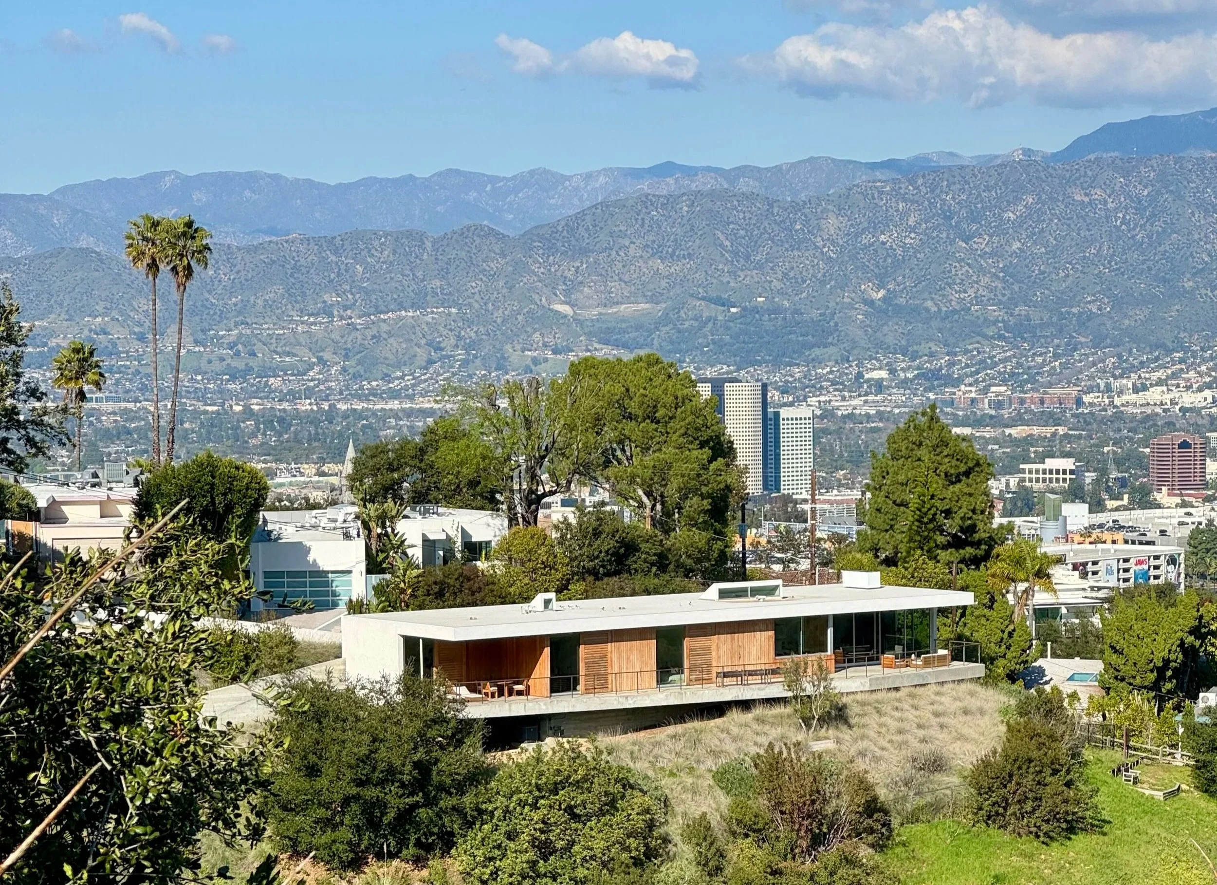 Cityscape view from Mulholland Drive, Studio City  lush green trees, and mountains in the background under a partly cloudy sky.