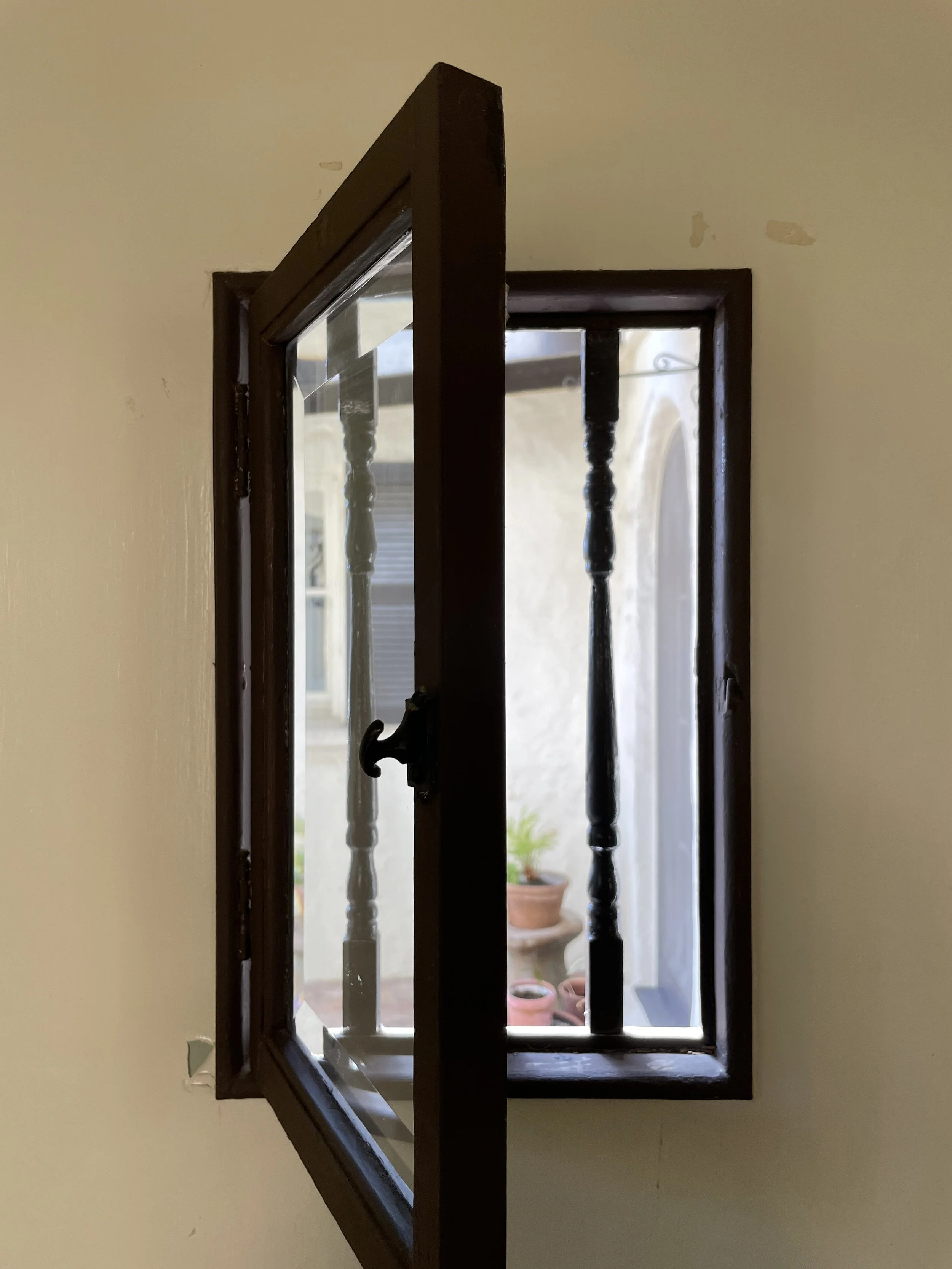 Open window with wooden frame and decorative balusters, located in a beige wall, revealing potted plants and building outside in Los Feliz neighborhood.