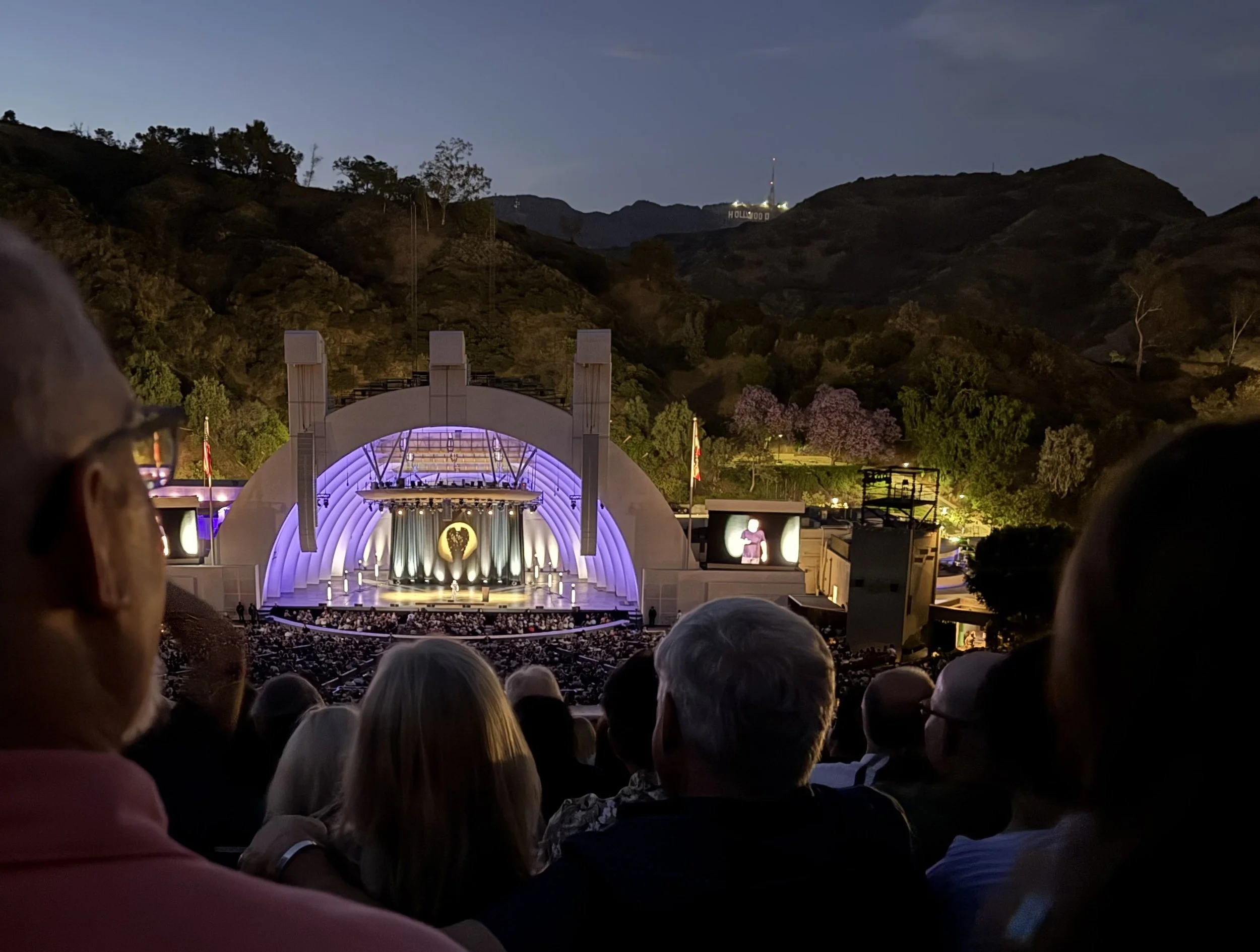 People seated at the Hollywood Bowl, an outdoor concert in the evening, with the Hollywood Sign visible on a hill in the background and mountains surrounding the area.