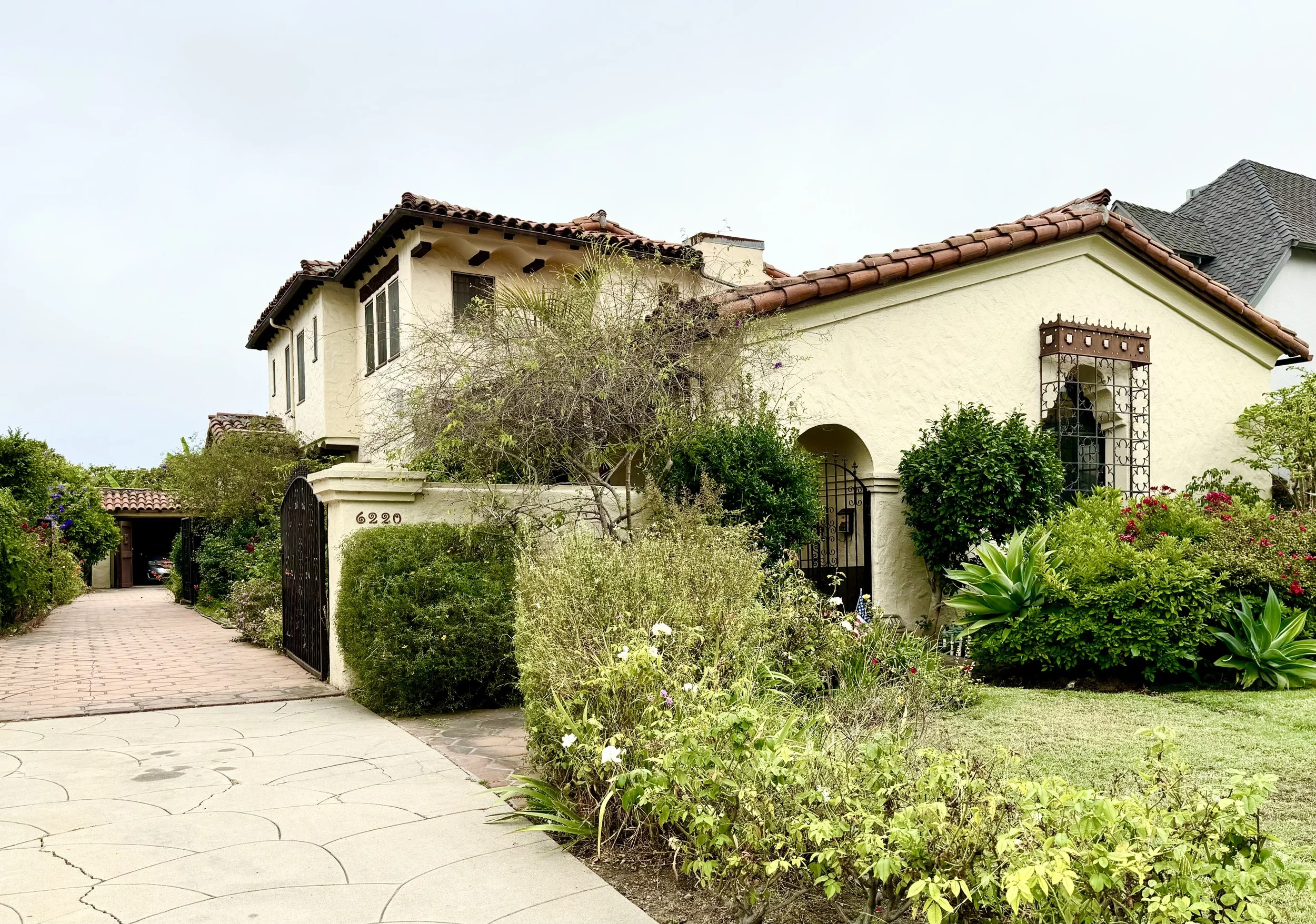 A large house with beige stucco walls, red clay tile roof, and a small balcony with black wrought iron railing. The front yard has various bushes and flowering plants, a paved walkway with cracks, and a black gate at the entrance. The house number 6220 is visible on the left side.
