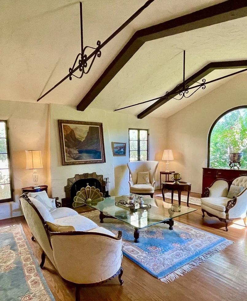 Living room in Brentwood, with vintage furniture, a glass coffee table, framed landscape paintings, and large windows with view of green trees.