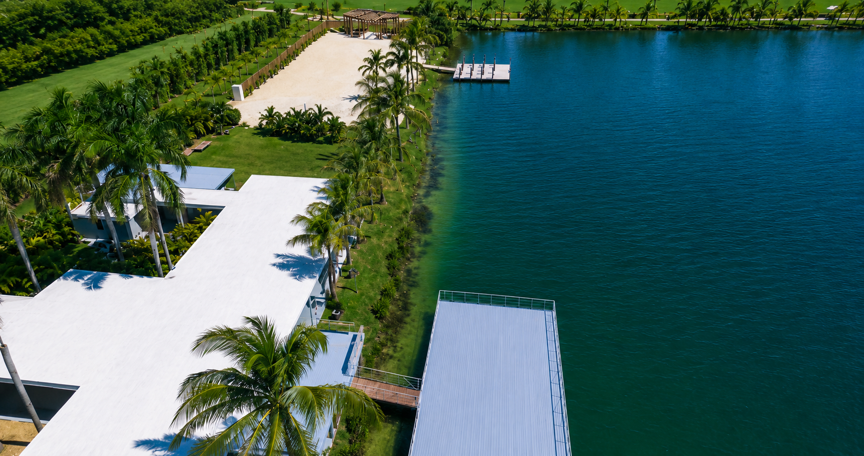 Aerial view of a lakeside resort with white buildings, palm trees, green lawn, sandy area, and a wooden dock extending into the blue water.