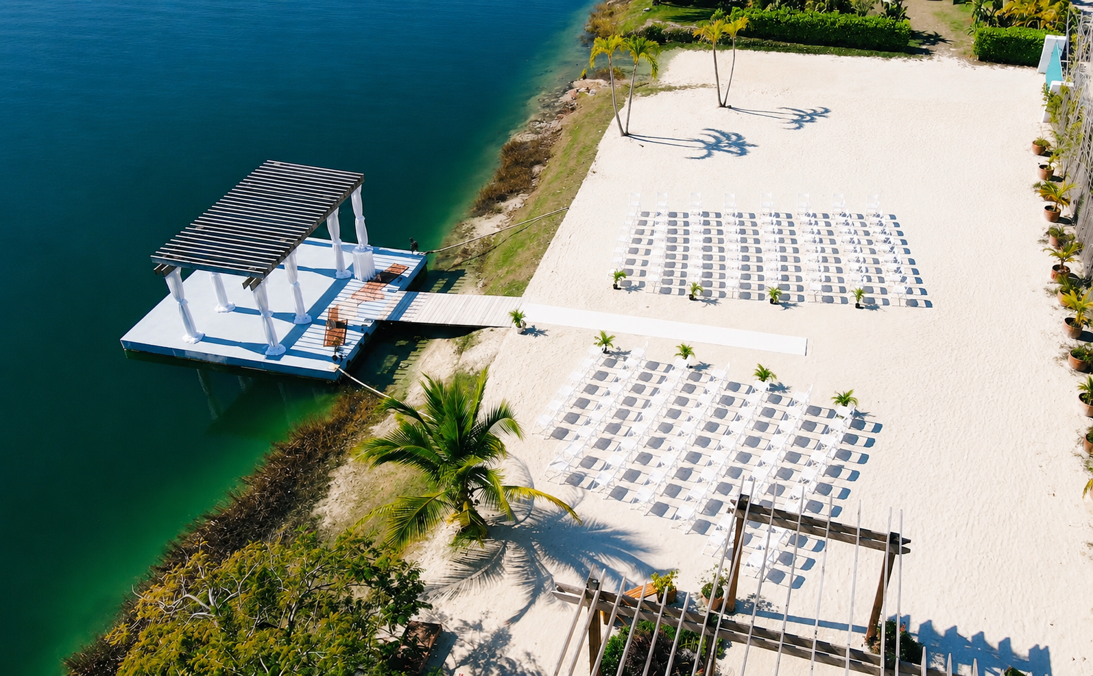 Aerial view of a beachside setup with white lounge chairs arranged on the sand, small plants lining the area, a wooden bridge, and a shaded pavilion over the water.