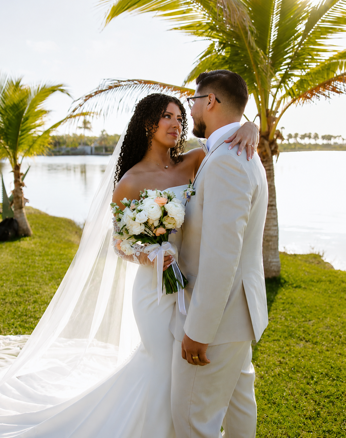 A bride and groom stand close by a water body, with palm trees in the background, during sunset. The bride holds a bouquet of white roses and wears a white wedding dress with a veil, while the groom wears a white tuxedo.