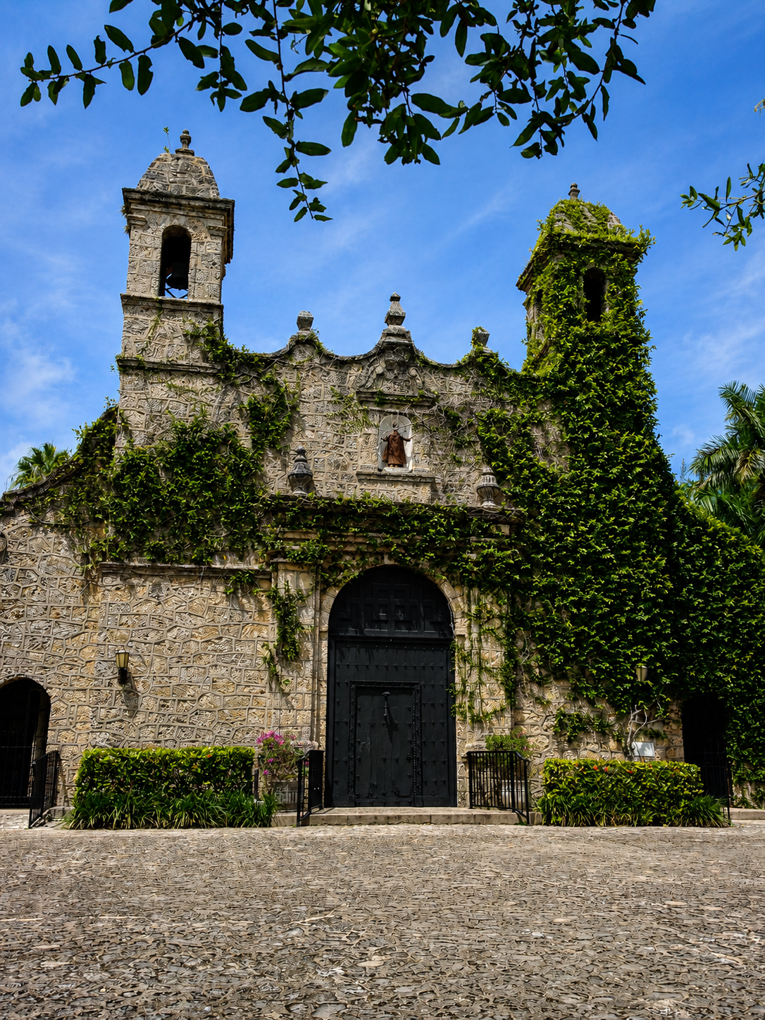 Historic stone church building with arches, towers, and clerestory windows, covered in green vines, with a black iron gate and cobblestone courtyard.