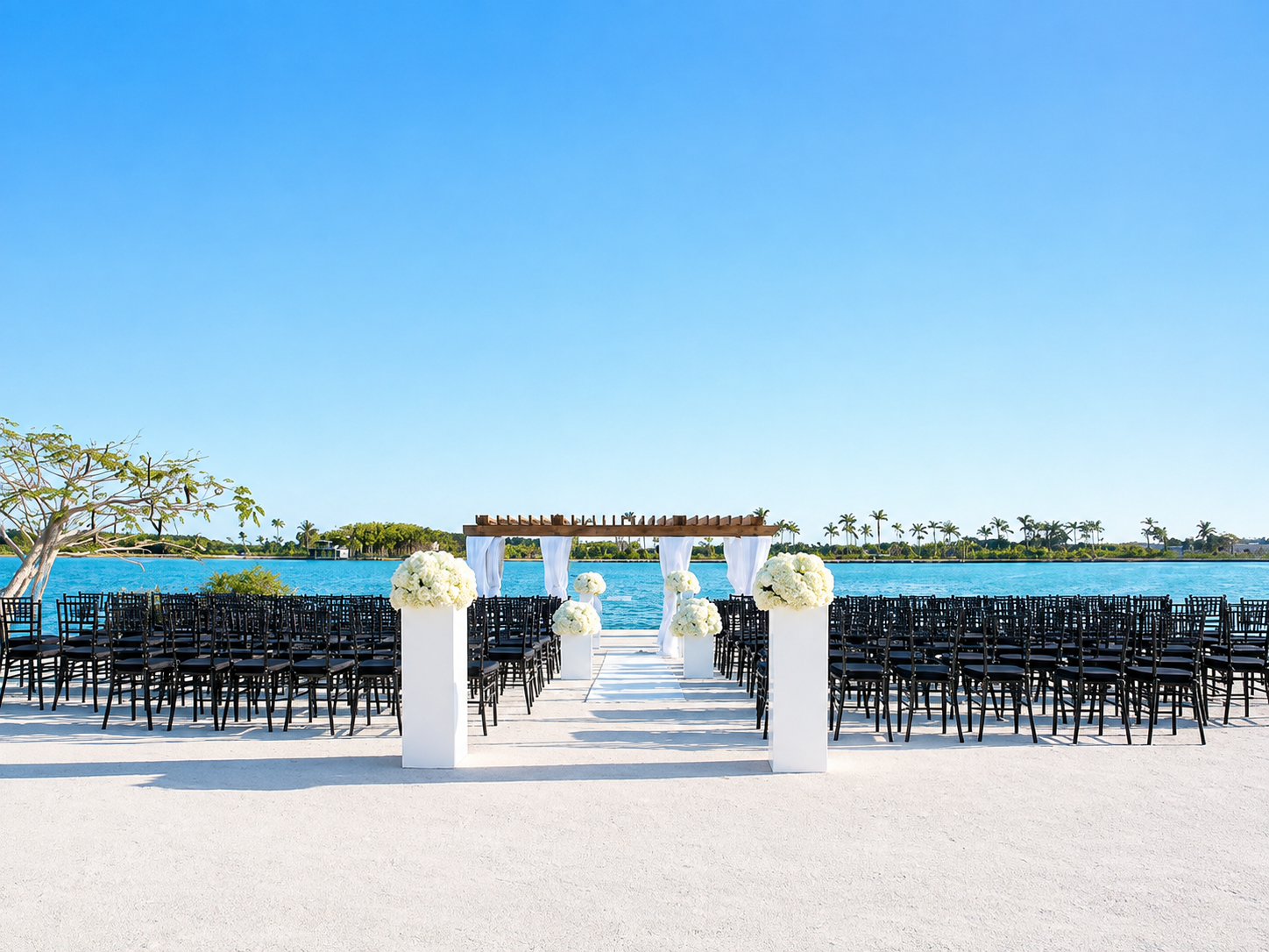 Outdoor wedding setup by a lake with black chairs, white floral arrangements on pillars, and a wooden arbor with white drapery against a clear blue sky.