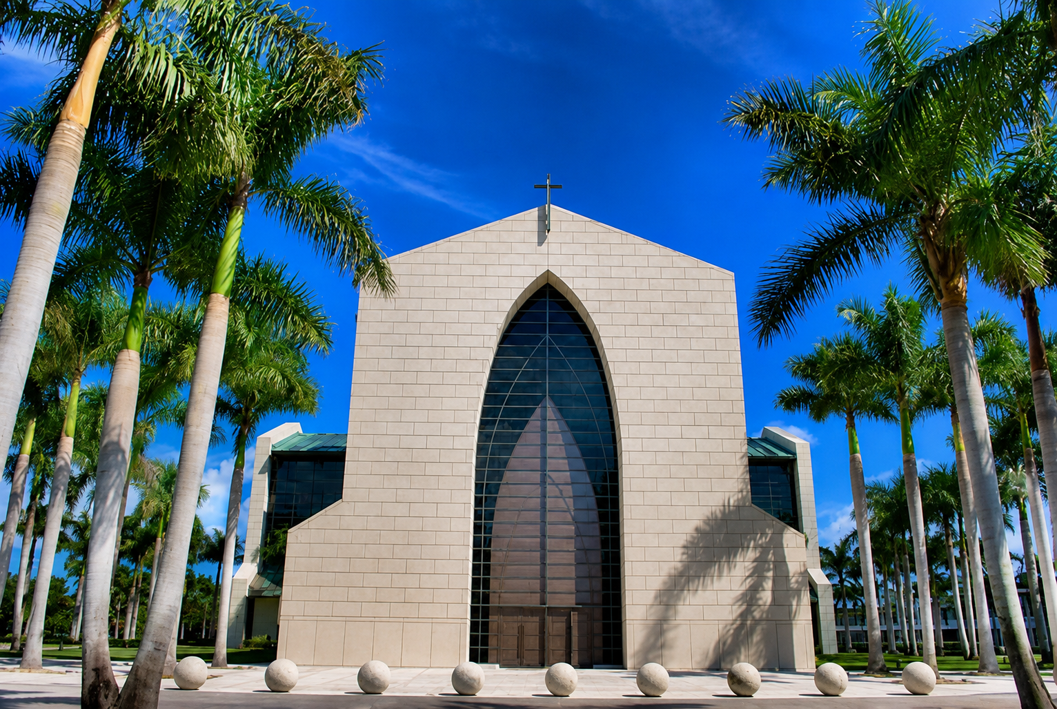 Modern church with a large cross on the roof, surrounded by palm trees, under a blue sky with some clouds.