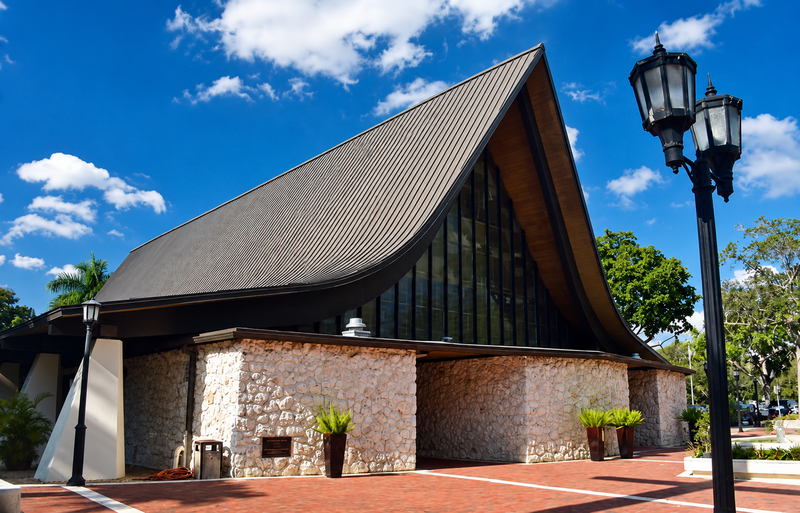 A modern building with a curved metal roof, stone walls, large glass windows, and a brick walkway, with a black street lamp and green trees in the background.