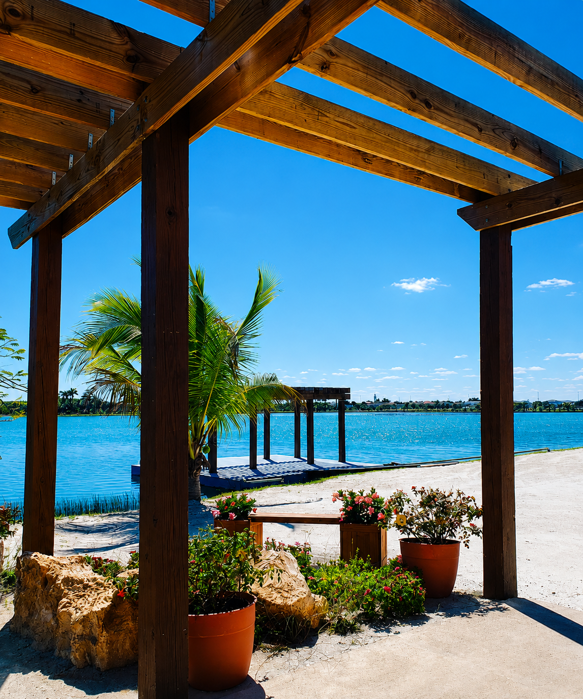 Beachside wooden pergola with potted plants, view of water, and palm trees under a bright blue sky.