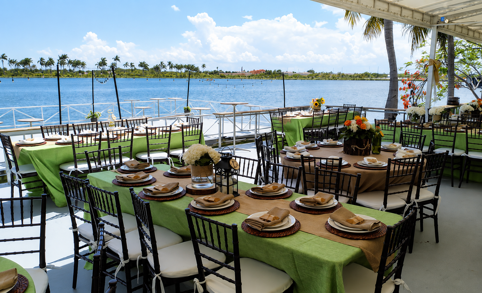 Dinner setup on a boat with tables covered in green and beige tablecloths, decorated with floral centerpieces, over a water body with palm trees and a partly cloudy sky in the background.
