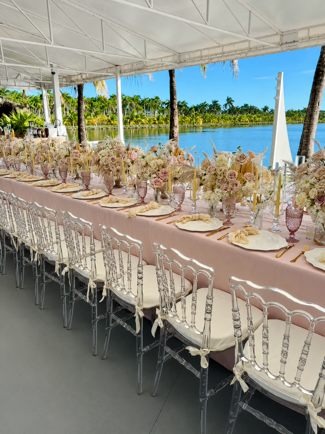 A long outdoor dining table set for a celebration under a white canopy, overlooking a river with palm trees in the background. The table is decorated with pink and cream floral arrangements, candles, and elegant plates and cutlery.