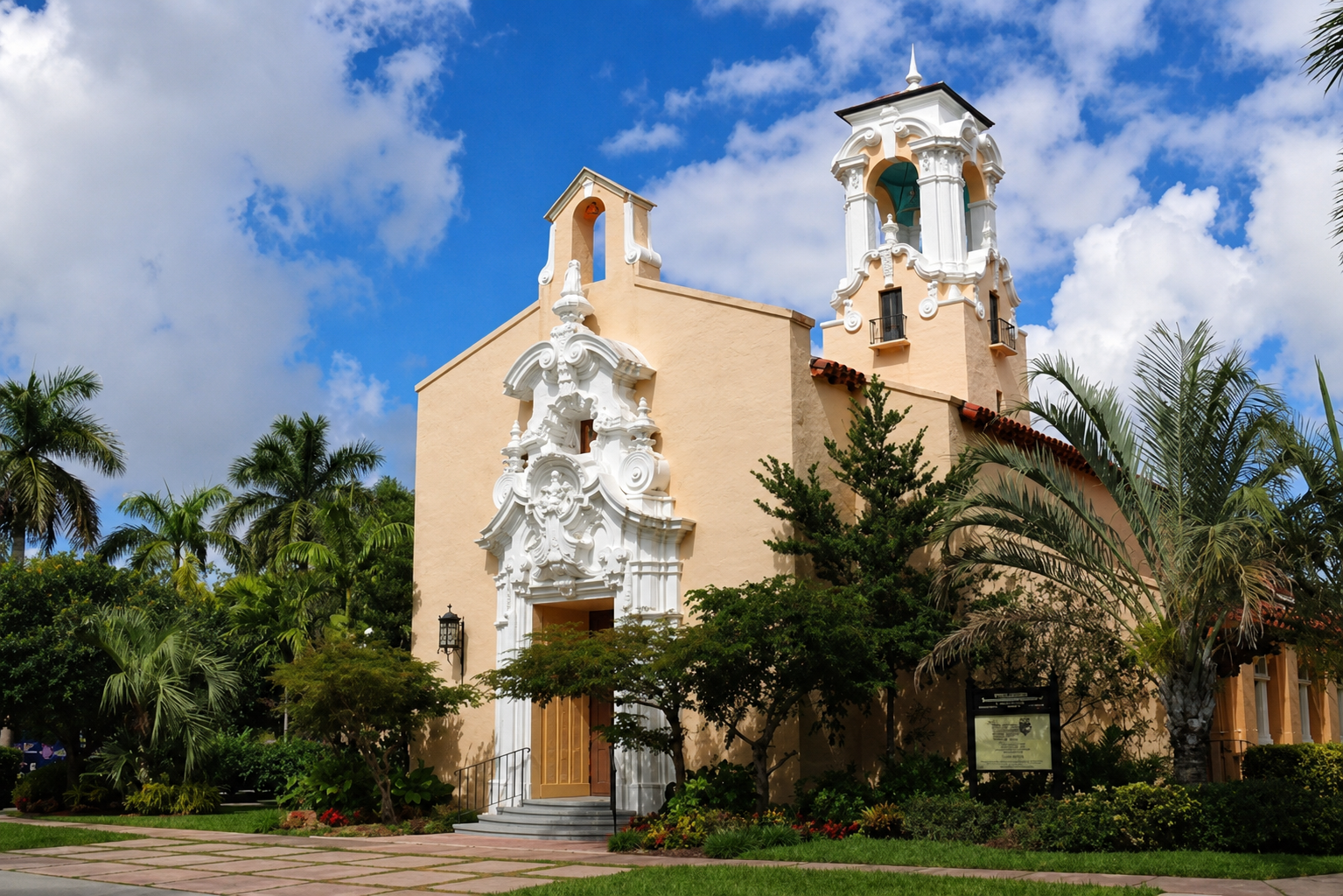 A Spanish-style church with stucco walls, decorative white trim, and a bell tower, surrounded by lush greenery and palm trees under a partly cloudy blue sky.