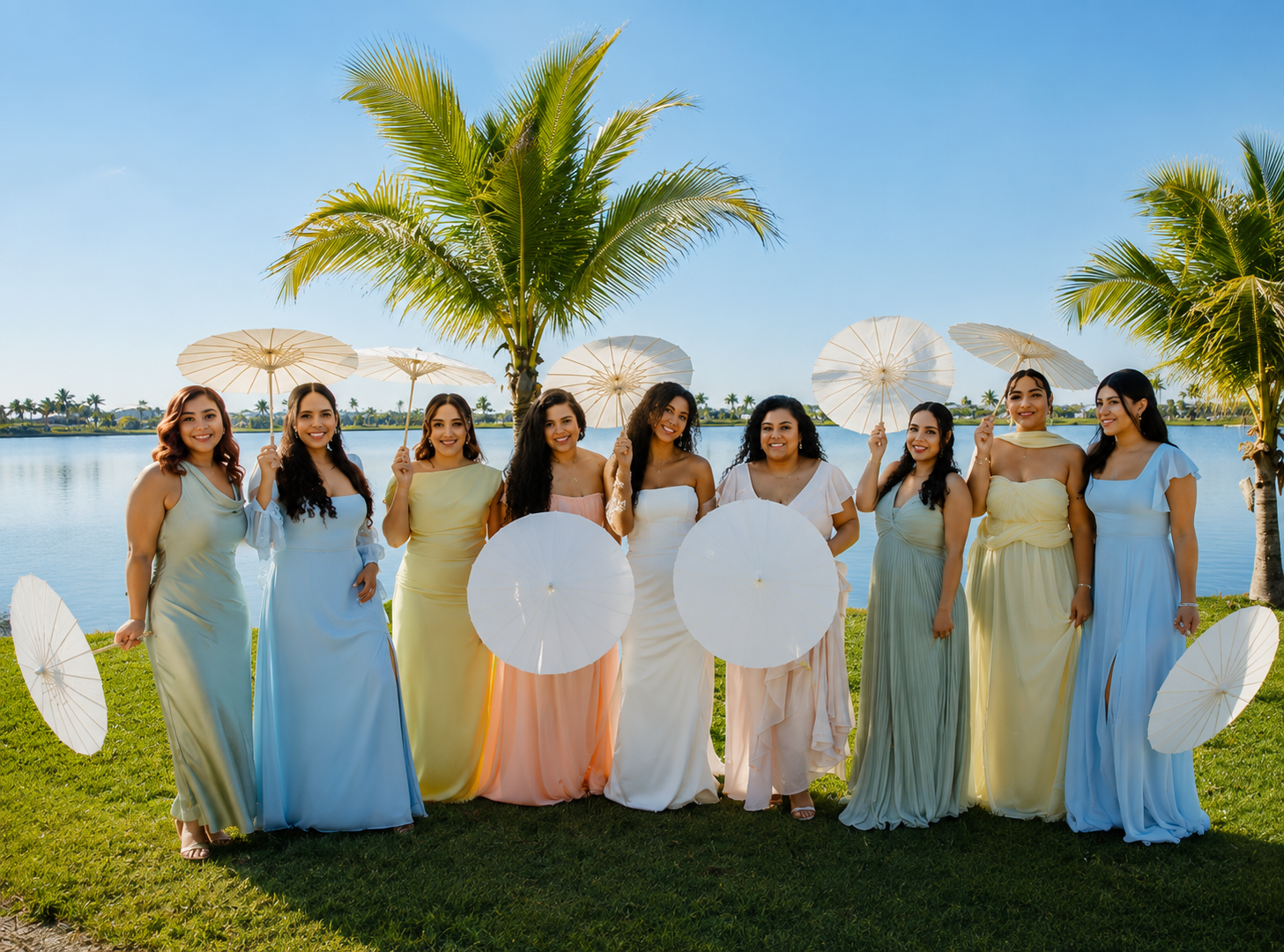 A group of nine women dressed in pastel-colored dresses standing on grass near a body of water, holding white parasols, with palm trees and a clear blue sky in the background.