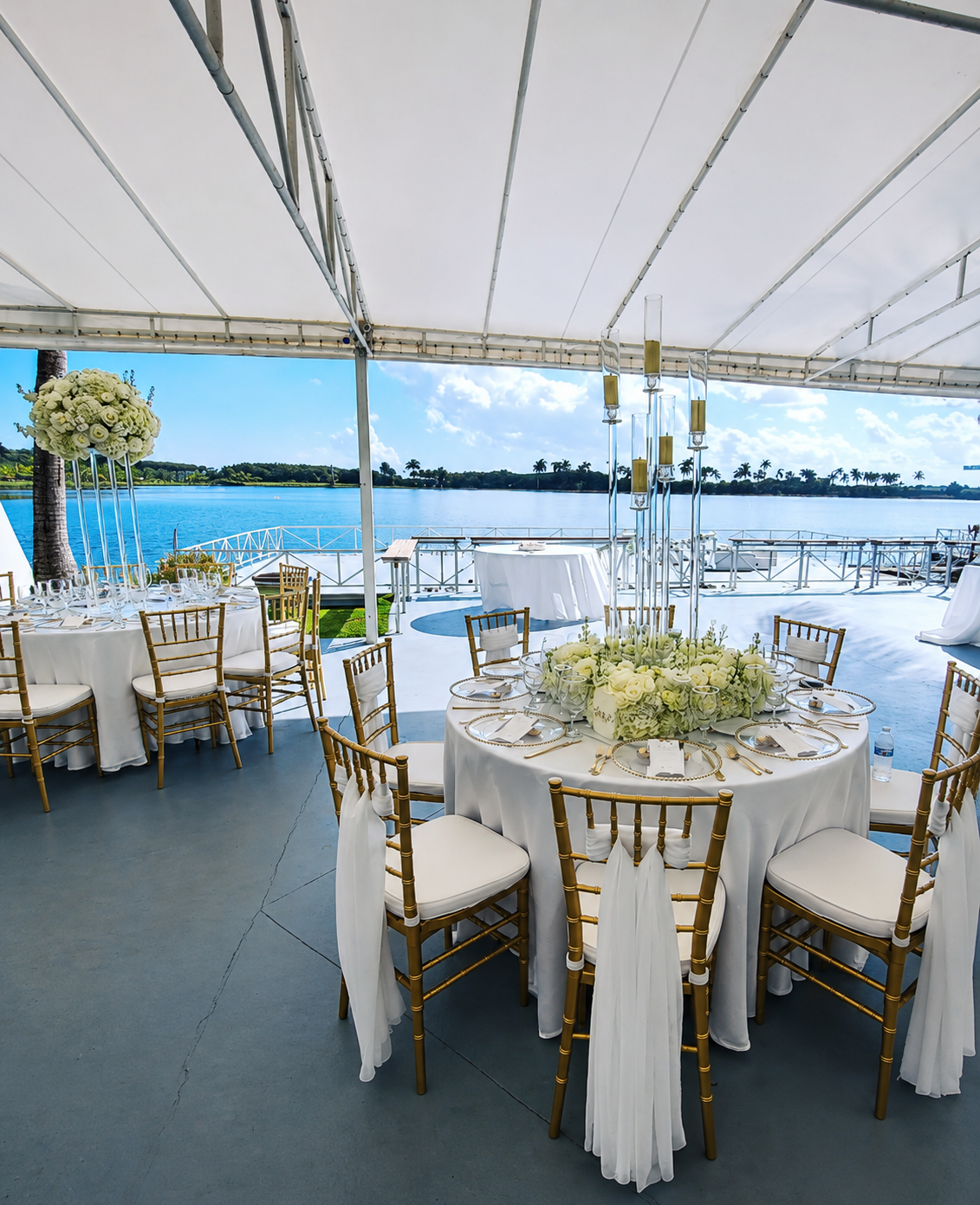 Elegant outdoor waterfront wedding reception setup with round tables, gold chairs, white tablecloths, floral centerpieces, and a scenic view of water and palm trees.