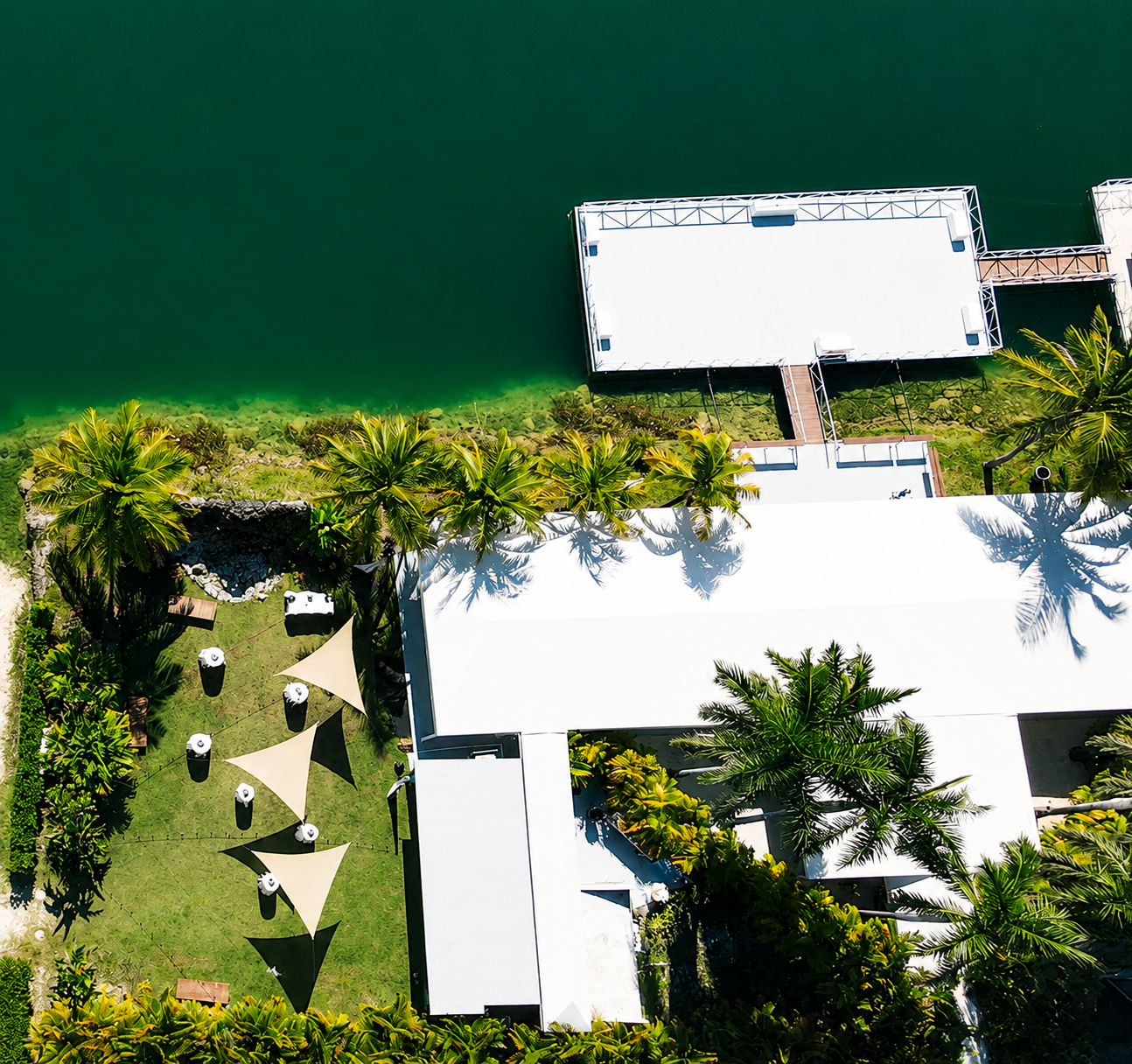 Aerial view of a tropical resort with a white building, palm trees, a grassy area with umbrellas, and a dock leading out to the water.