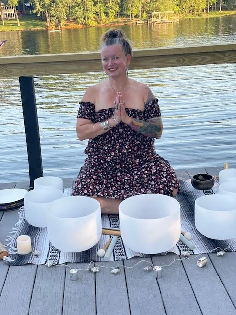 Shannon Somogyi sitting cross-legged on a dock by the water, surrounded by singing bowls and other meditation tools, during sound bath at Lake Gaston