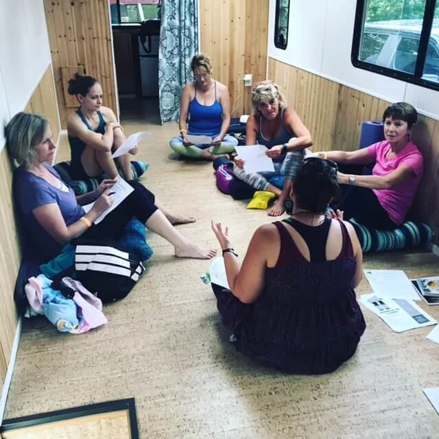 Six women sitting in a circle on a carpeted floor reading papers and participating in a discussion with Shannon during a wellness workshop.