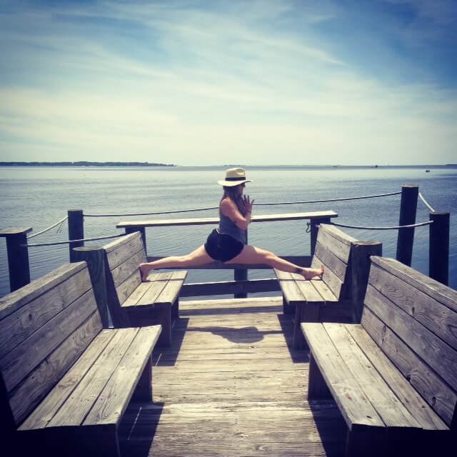 A woman practicing yoga in a split pose on a wooden pier overlooking a calm body of water, with blue sky and distant land visible.