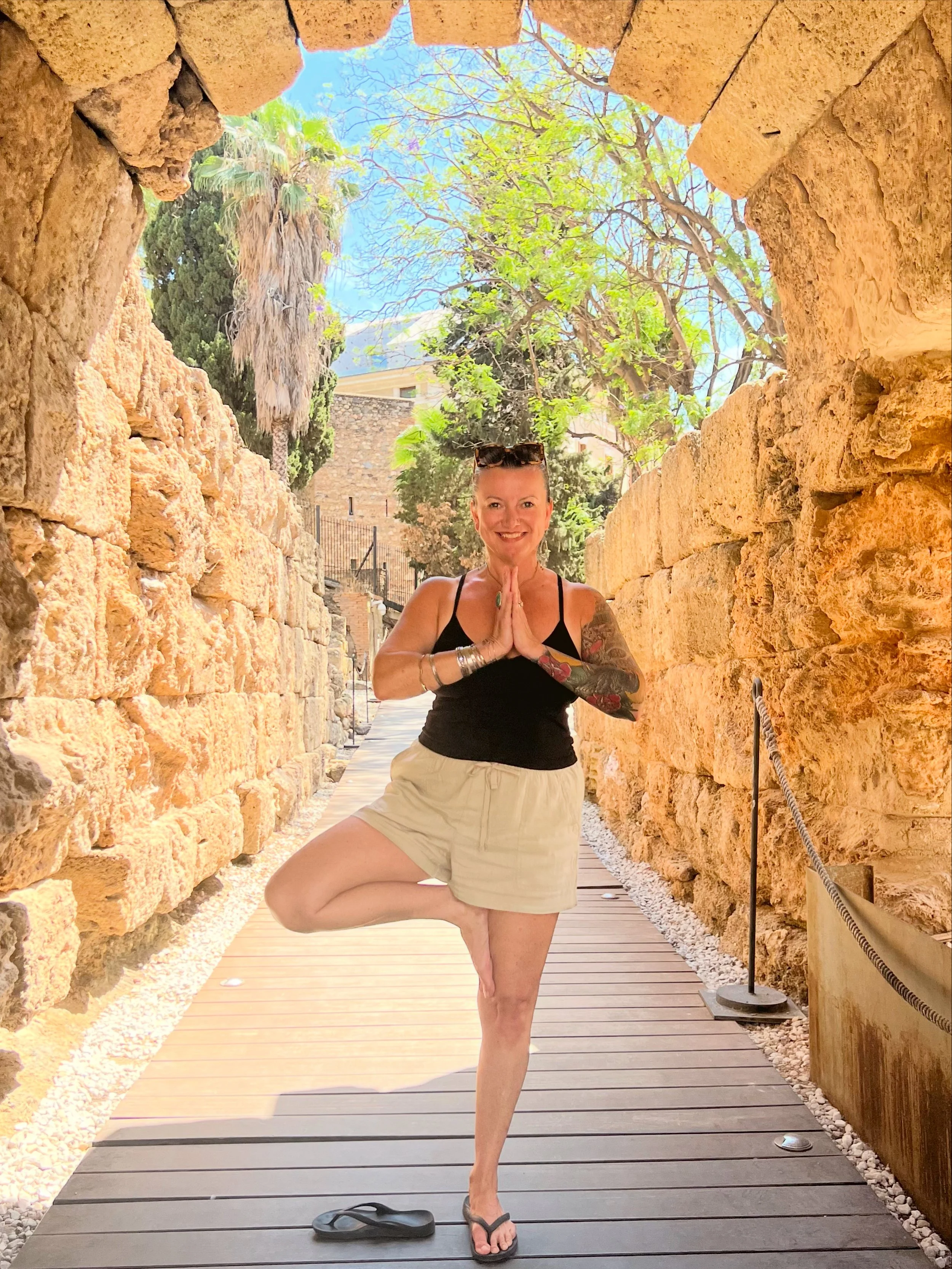 Shannon Somogyi practicing yoga in a peaceful outdoor setting, standing on a wooden pathway between stone walls, with trees, a blue sky, and palm trees in the background.