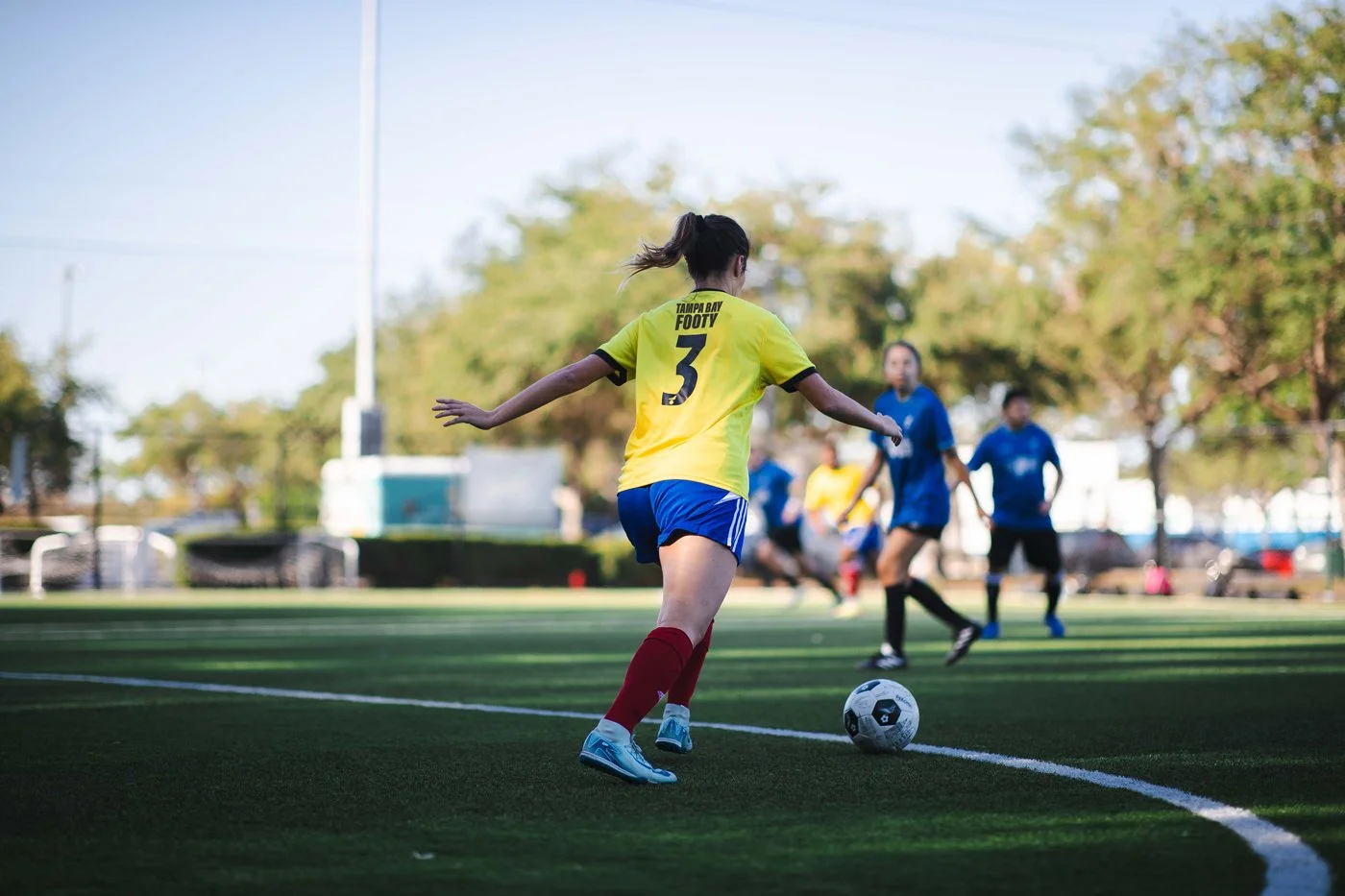 A Tampa Bay Footy female soccer player in a yellow jersey and blue shorts preparing to kick a soccer ball during a match on a green field, with other players and trees in the background.