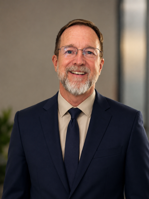 A smiling middle-aged man with glasses, a beard, wearing a navy suit, white shirt, and navy tie, standing indoors with a blurred background.