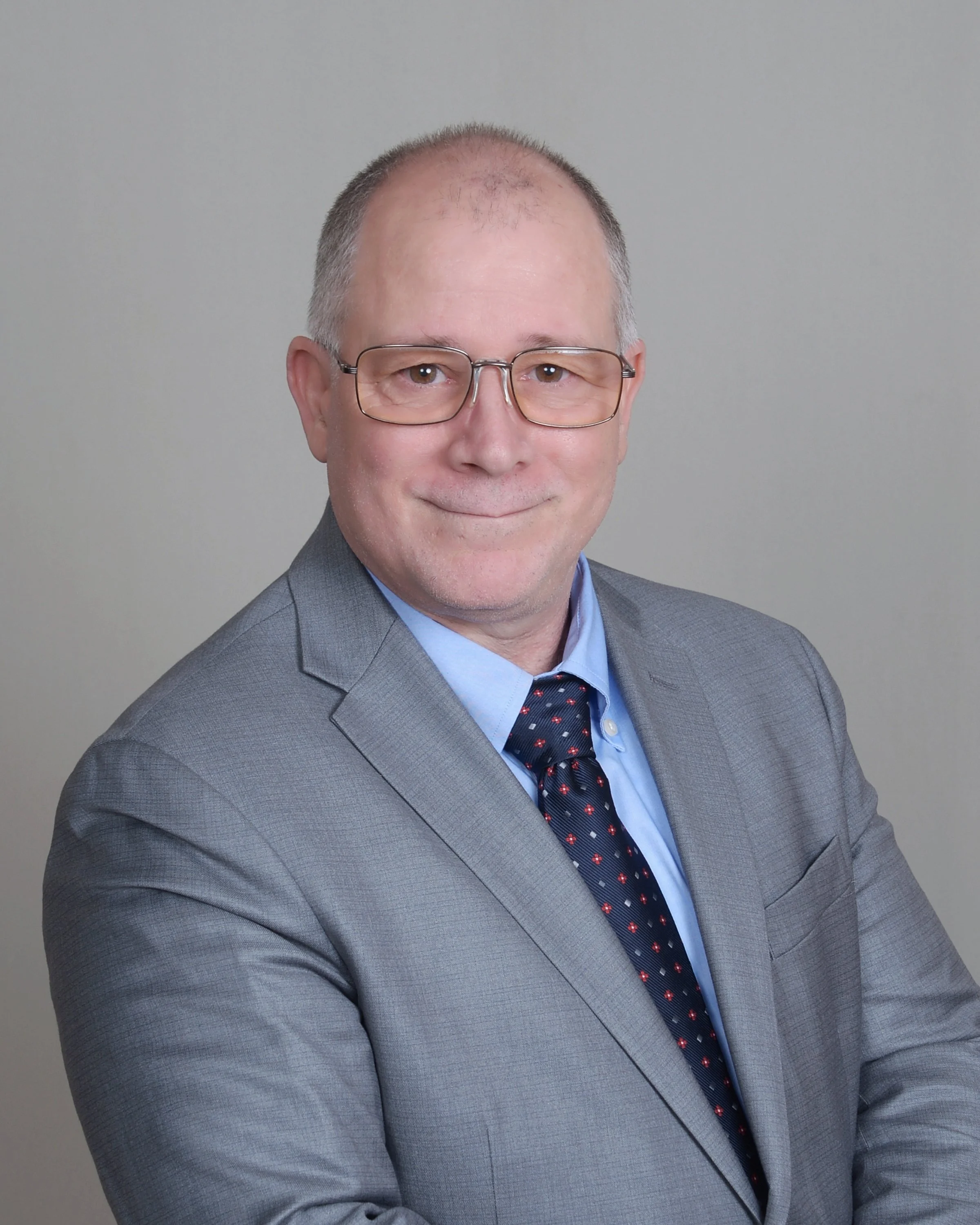 A middle-aged man with short gray hair, glasses, and light skin, dressed in a gray suit, light blue shirt, and a dark tie with small red and white patterns, smiling at the camera against a plain gray background.