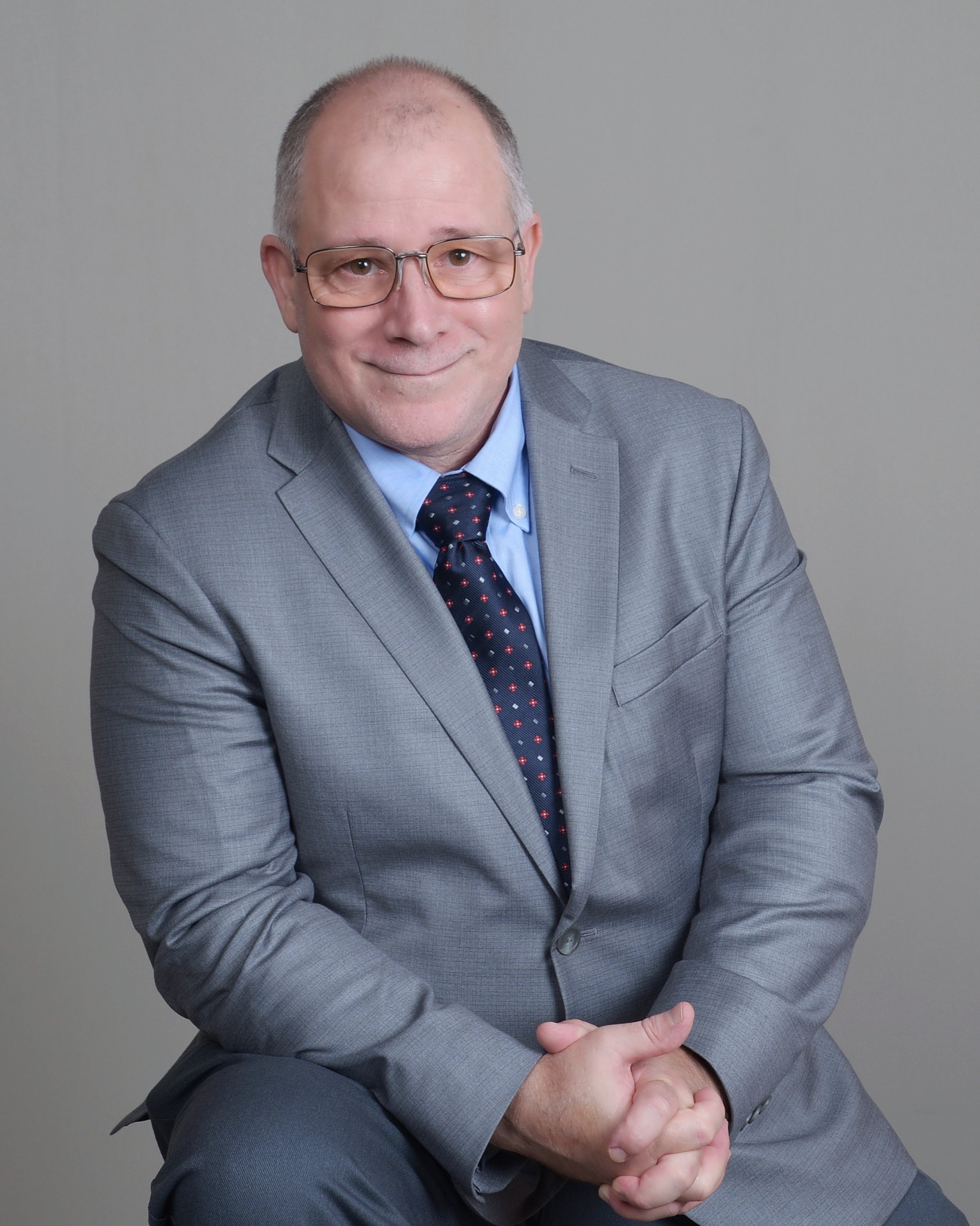 Man wearing glasses and a gray suit with a light blue shirt and dark patterned tie, posing with his hands clasped and smiling against a plain gray background.